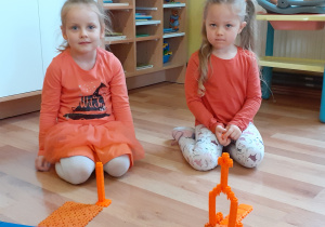2 dziewczynki pokazuje zbudowane przez siebie konstrukcje z pomarańczowych klocków.
